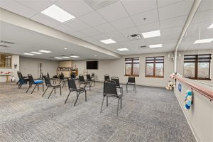 Spacious group activity and exercise room featuring widely spaced chairs, a wall mirror with a handrail, and large windows at Cedar Crest Senior Living, a 55+ retirement community in Janesville, WI.